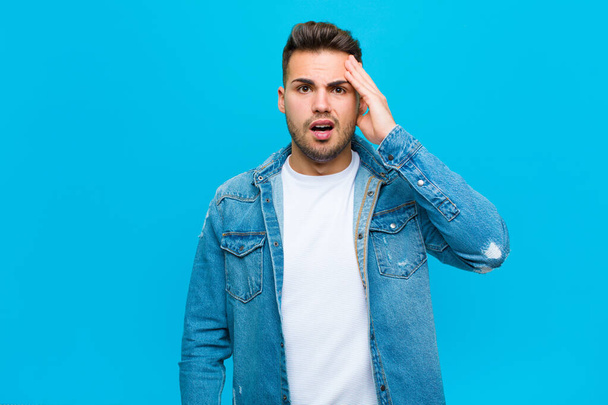 young hispanic man looking happy, astonished and surprised, smiling and realizing amazing and incredible good news against blue background - Foto, imagen