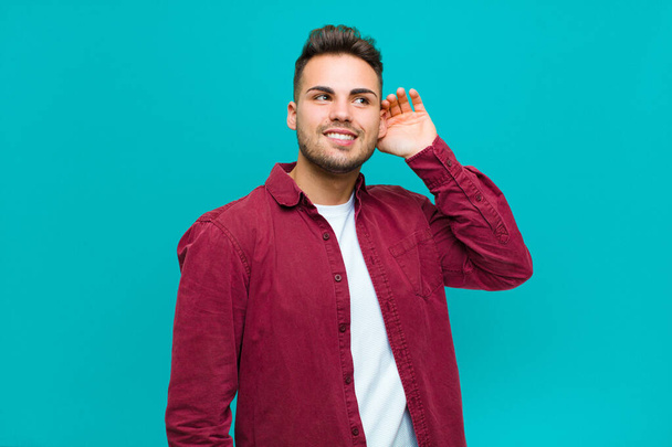 young hispanic man smiling, looking curiously to the side, trying to listen to gossip or overhearing a secret against blue background - Foto, imagen