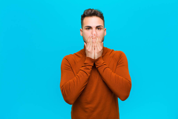 young hispanic man happy and excited, surprised and amazed covering mouth with hands, giggling with a cute expression against blue background - Φωτογραφία, εικόνα