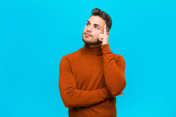 young hispanic man with a concentrated look, wondering with a doubtful expression, looking up and to the side against blue background - Foto, afbeelding