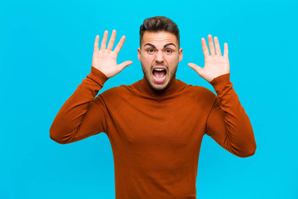 young hispanic man screaming in panic or anger, shocked, terrified or furious, with hands next to head against blue background - Foto, imagen
