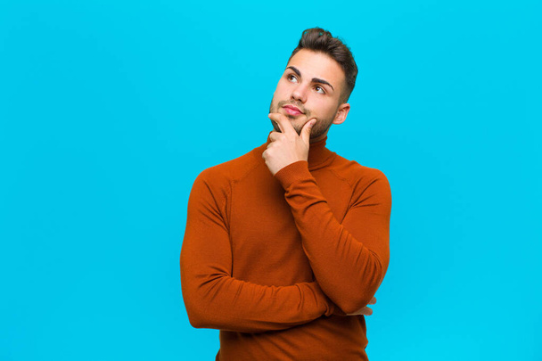 young hispanic man feeling thoughtful, wondering or imagining ideas, daydreaming and looking up to copy space against blue background - Photo, Image