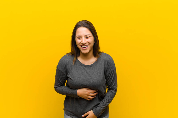 latin american woman laughing out loud at some hilarious joke, feeling happy and cheerful, having fun isolated against yellow wall - Valokuva, kuva