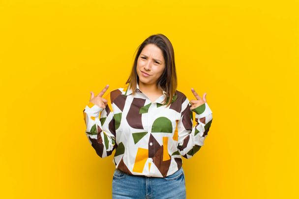 Latinskoamerická žena se špatným postojem vypadá hrdě a agresivně, ukazuje nahoru nebo dělá zábavné znamení s rukama izolovanýma o žlutou stěnu - Fotografie, Obrázek