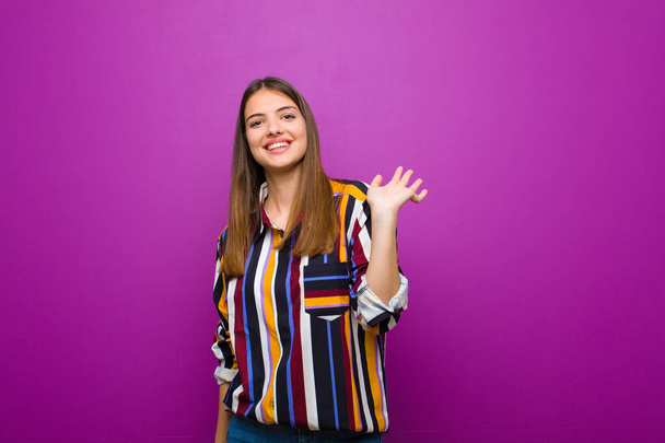 young pretty woman smiling happily and cheerfully, waving hand, welcoming and greeting you, or saying goodbye against purple background - Foto, Bild