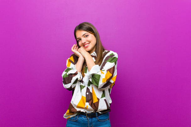 young pretty woman feeling in love and looking cute, adorable and happy, smiling romantically with hands next to face against purple background - 写真・画像