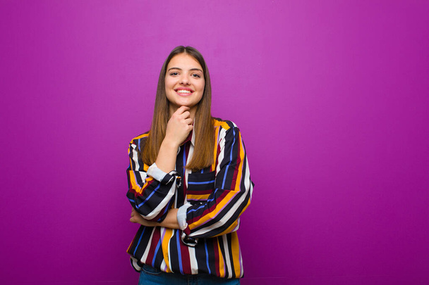 young pretty woman smiling, enjoying life, feeling happy, friendly, satisfied and carefree with hand on chin against purple background - Zdjęcie, obraz
