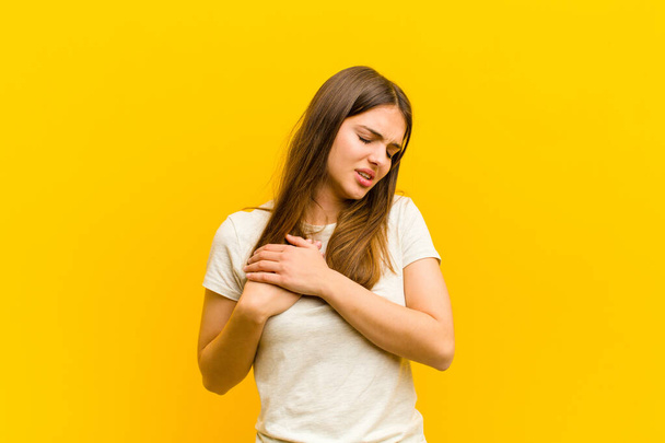 young pretty woman looking sad, hurt and heartbroken, holding both hands close to heart, crying and feeling depressed against orange background - Photo, Image