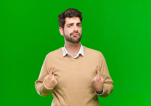 young businessman with a bad attitude looking proud and aggressive, pointing upwards or making fun sign with hands against green background - Foto, imagen