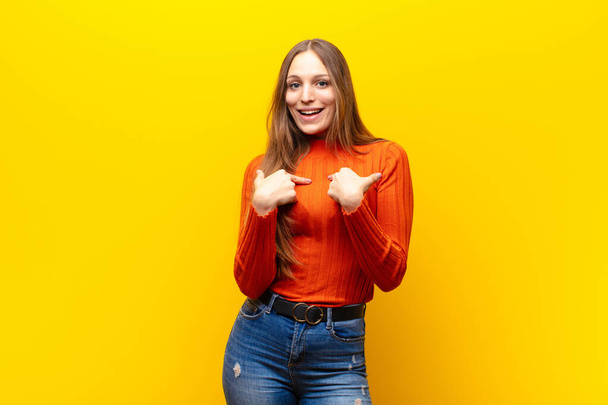 young pretty woman feeling happy, surprised and proud, pointing to self with an excited, amazed look against orange background - Foto, imagen