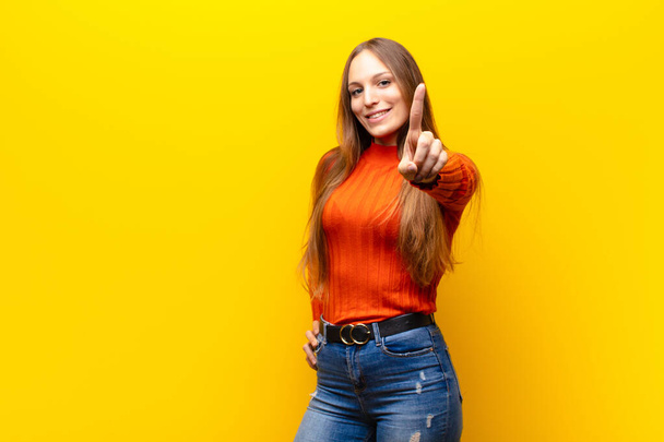 young pretty woman smiling proudly and confidently making number one pose triumphantly, feeling like a leader against orange background - Фото, изображение
