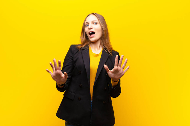 young pretty businesswoman feeling stupefied and scared, fearing something frightening, with hands open up front saying stay away against orange background - Фото, зображення