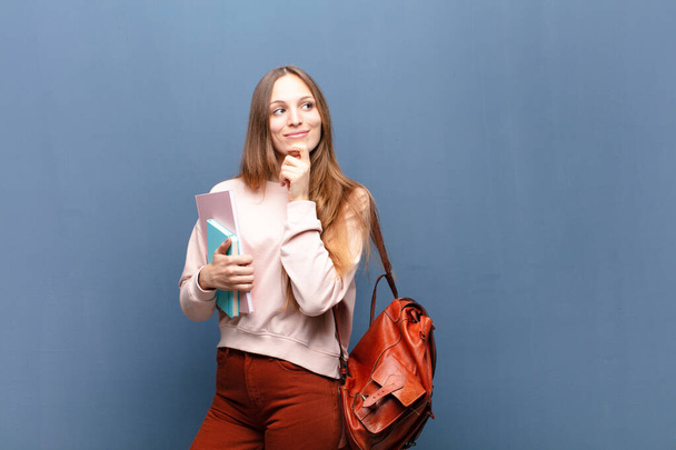 young pretty student woman with books and bag against blue wall with a copy space - Фото, изображение