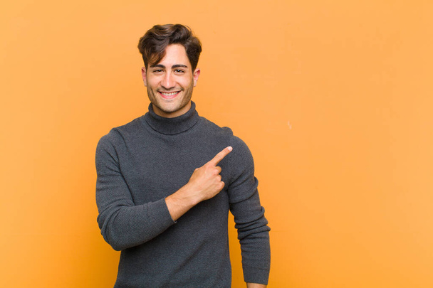young handsome man smiling cheerfully, feeling happy and pointing to the side and upwards, showing object in copy space against orange background - Фото, изображение