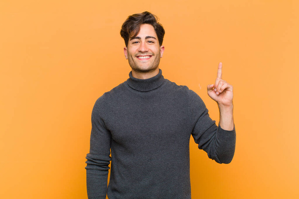 young handsome man smiling cheerfully and happily, pointing upwards with one hand to copy space against orange background - Foto, Imagen