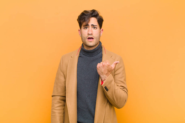 young handsome man looking astonished in disbelief, pointing at object on the side and saying wow, unbelievable against orange background - Photo, image