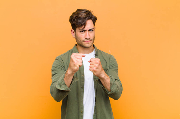 young handsome man looking confident, angry, strong and aggressive, with fists ready to fight in boxing position against orange background - Φωτογραφία, εικόνα
