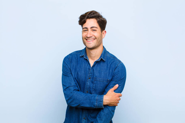junger gutaussehender Mann, der schüchtern und fröhlich lacht, mit einer freundlichen und positiven, aber unsicheren Haltung vor blauem Hintergrund - Foto, Bild