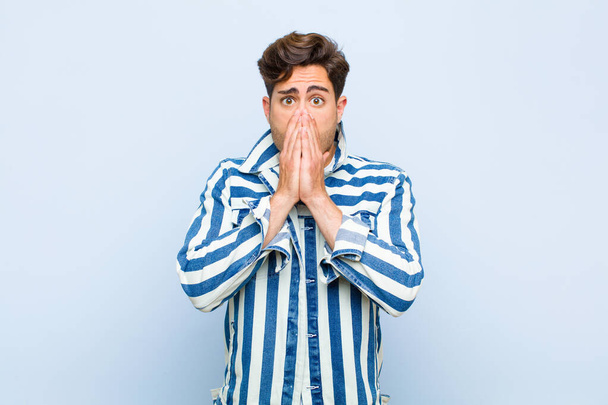 young handsome man feeling worried, upset and scared, covering mouth with hands, looking anxious and having messed up against blue background - Foto, Imagem