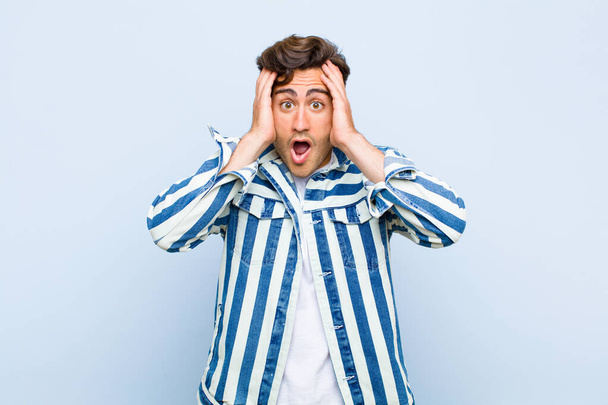 young handsome man looking unpleasantly shocked, scared or worried, mouth wide open and covering both ears with hands against blue background - Foto, Bild