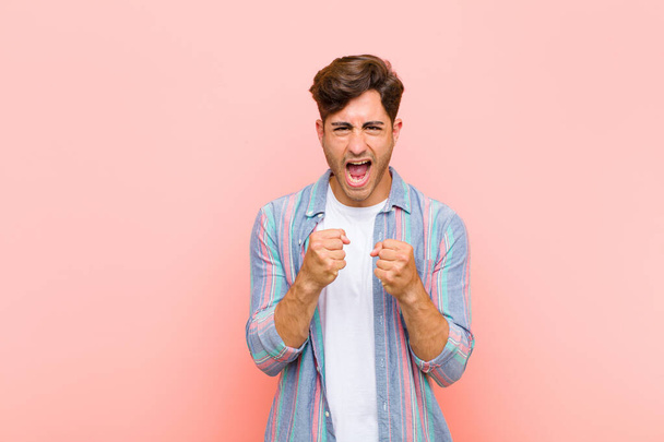 young handsome man shouting aggressively with annoyed, frustrated, angry look and tight fists, feeling furious against pink background - Photo, Image