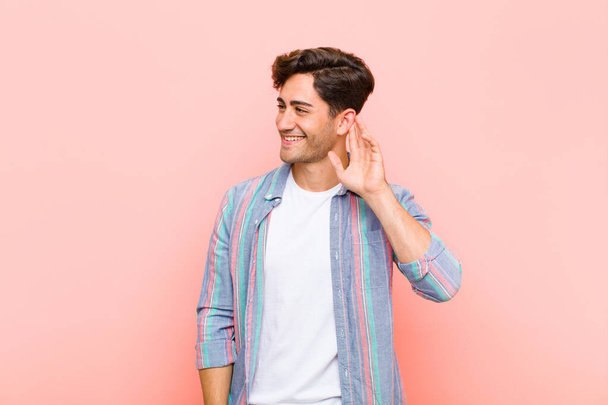 young handsome man smiling, looking curiously to the side, trying to listen to gossip or overhearing a secret against pink background - Photo, Image