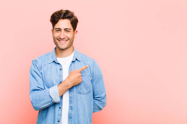 young handsome man smiling cheerfully, feeling happy and pointing to the side and upwards, showing object in copy space against pink background - Fotó, kép