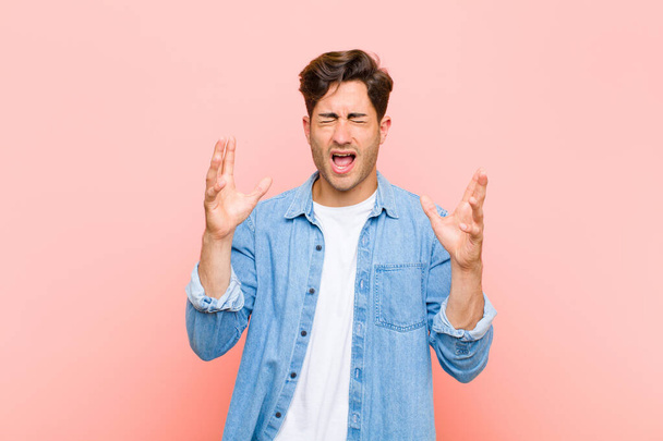 young handsome man furiously screaming, feeling stressed and annoyed with hands up in the air saying why me against pink background - Fotoğraf, Görsel