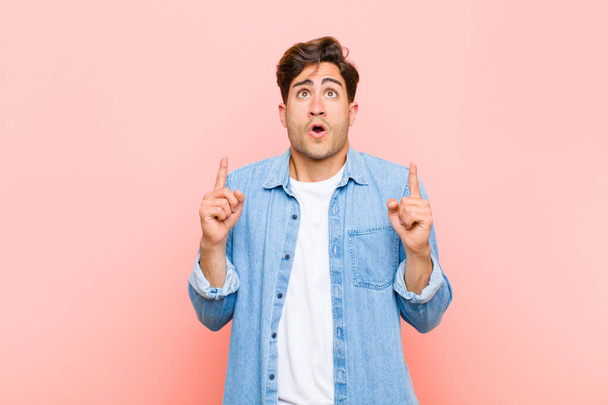 young handsome man feeling awed and open mouthed pointing upwards with a shocked and surprised look against pink background - Foto, imagen