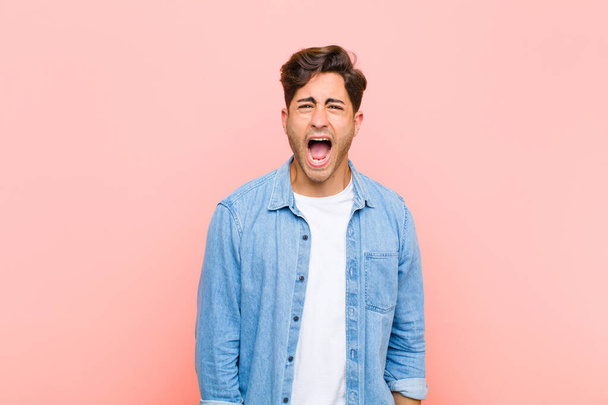 young handsome man shouting aggressively, looking very angry, frustrated, outraged or annoyed, screaming no against pink background - Фото, изображение