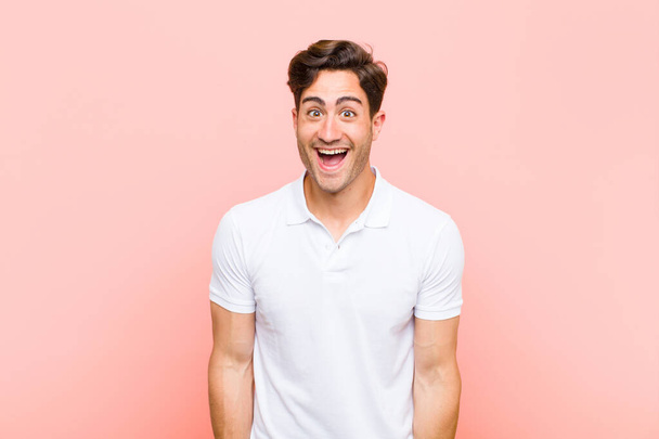 young handsome man looking happy and pleasantly surprised, excited with a fascinated and shocked expression against pink background - Photo, Image