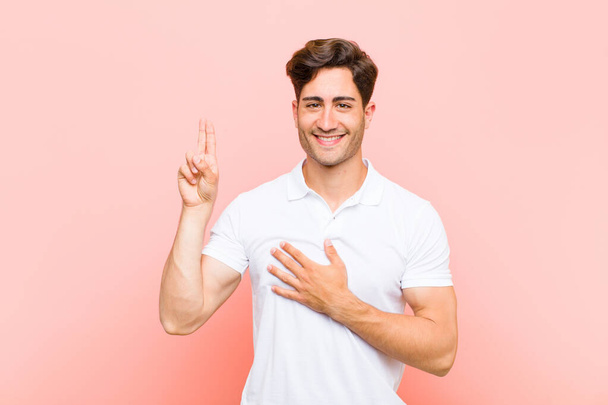 young handsome man looking happy, confident and trustworthy, smiling and showing victory sign, with a positive attitude against pink background - Foto, Bild