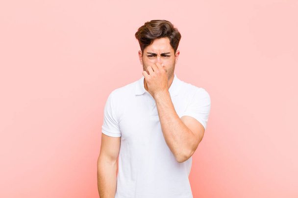 young handsome man feeling disgusted, holding nose to avoid smelling a foul and unpleasant stench against pink background - Photo, Image