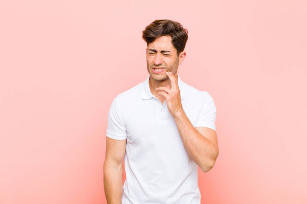 young handsome man holding cheek and suffering painful toothache, feeling ill, miserable and unhappy, looking for a dentist against pink background - Fotó, kép