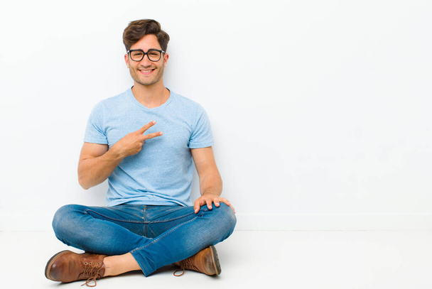 young handsome man feeling happy, positive and successful, with hand making v shape over chest, showing victory or peace sitting on the floor - Foto, imagen