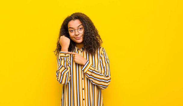 young pretty african american woman looking impatient and angry, pointing at watch, asking for punctuality, wants to be on time against yellow wall - Photo, image