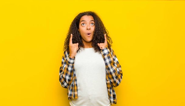 young pretty african american woman looking shocked, amazed and open mouthed, pointing upwards with both hands to copy space against yellow wall - Foto, Imagem