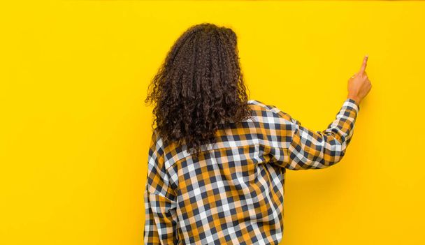 young pretty african american woman standing and pointing to object on copy space, rear view against yellow wall - Zdjęcie, obraz