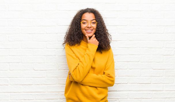 молодая черная женщина улыбается, наслаждаясь жизнью, чувствуя себя счастливым, дружелюбным, удовлетворенным и беззаботным с рукой на подбородке к кирпичной стене
 - Фото, изображение