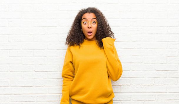 young black woman looking astonished in disbelief, pointing at object on the side and saying wow, unbelievable against brick wall - Photo, Image
