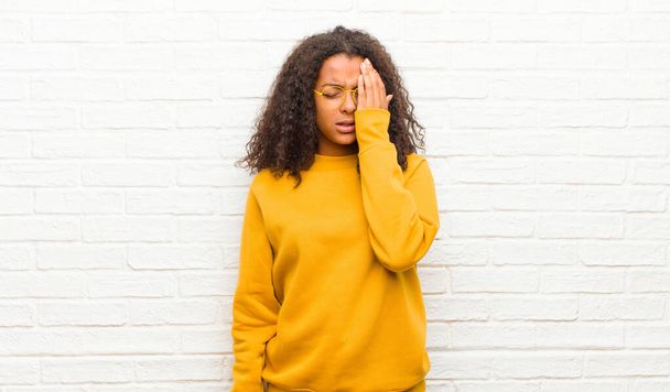 young black woman looking sleepy, bored and yawning, with a headache and one hand covering half the face against brick wall - Фото, изображение