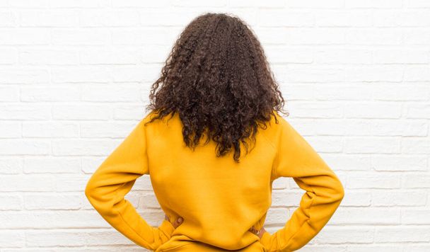 young black woman feeling confused or full or doubts and questions, wondering, with hands on hips, rear view against brick wall - Photo, Image