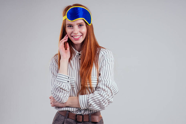 Good Morning. Smiling redhead young business woman in a striped shirt and blue yellow sleep mask white background studio - Foto, Imagen