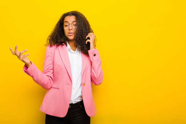 black business woman shrugging with a dumb, crazy, confused, puzzled expression, feeling annoyed and clueless against orange wall - Zdjęcie, obraz