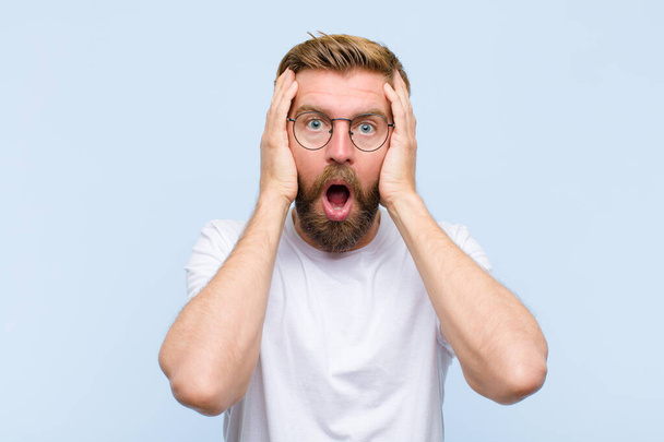young blonde adult man looking unpleasantly shocked, scared or worried, mouth wide open and covering both ears with hands - Foto, imagen