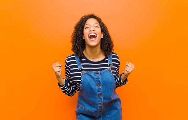 young pretty black woman feeling shocked, excited and happy, laughing and celebrating success, saying wow! against orange wall - Photo, image