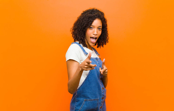 young pretty black woman feeling happy, cool, satisfied, relaxed and successful, pointing at camera, choosing you against orange wall - Foto, Bild