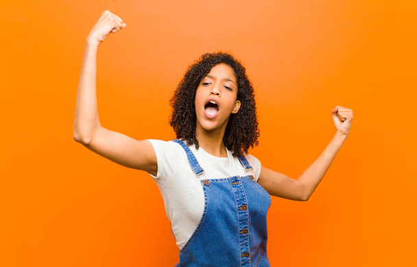 молодая красивая черная женщина, триумфально кричащая, выглядящая взволнованной, счастливой и удивленной победительницей, празднующей у оранжевой стены
 - Фото, изображение