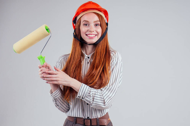 Smiling redhead businesswoman in a striped shirt builder worker holding a painting roller portrait on white studio - Foto, Imagen