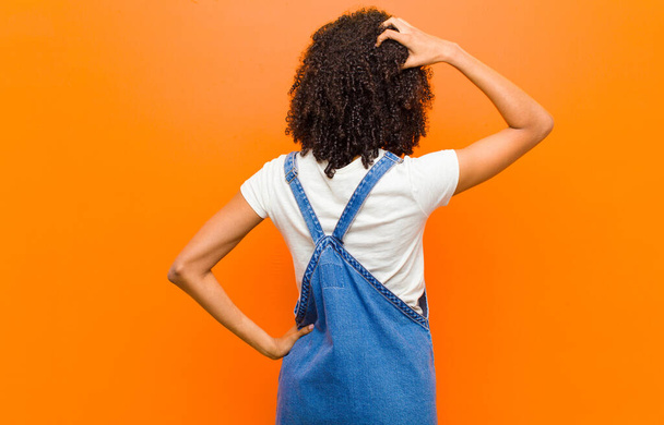 young pretty black woman feeling clueless and confused, thinking a solution, with hand on hip and other on head, rear view against orange wall - Fotografie, Obrázek
